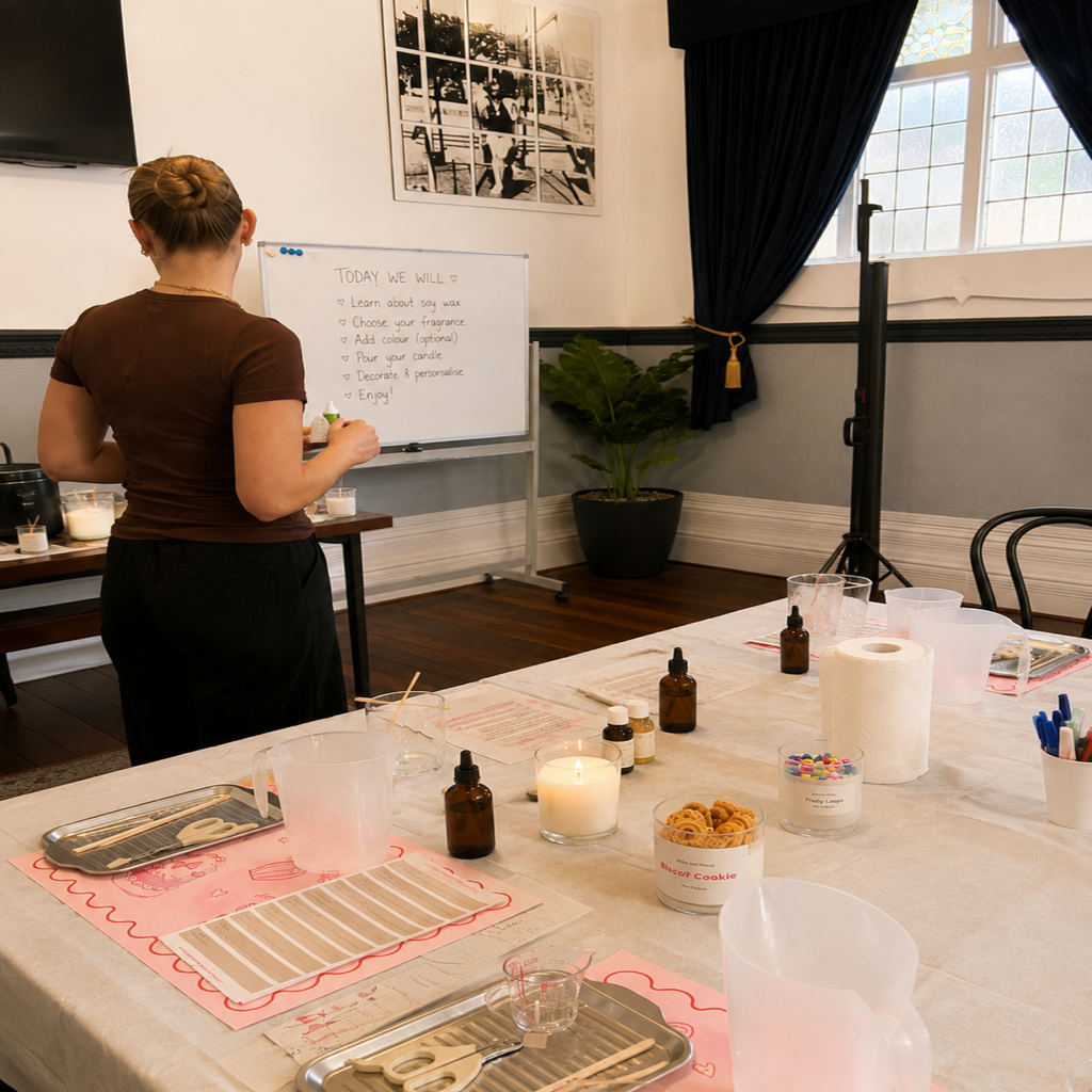A woman stands at a whiteboard in a workshop or studio setting, with a table in the foreground holding candles, amber bottles, plastic containers, and pink printed sheets.